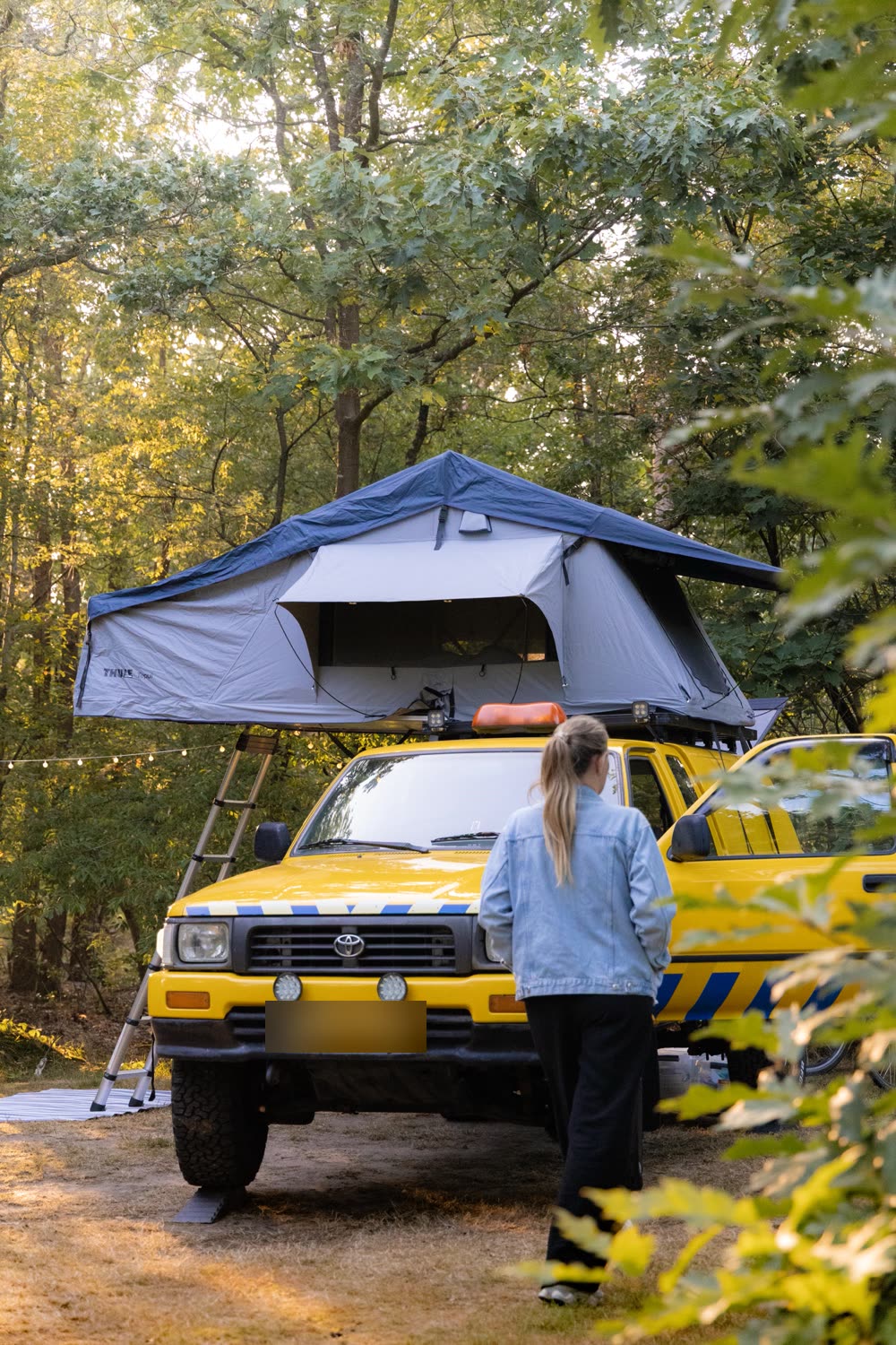 De gele Toyota Hilux met daktent op een kampeerplek in het bos, met Sharon ernaast